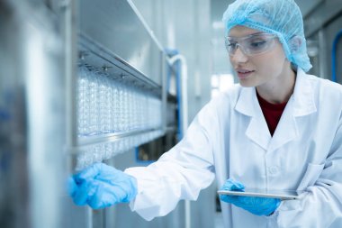 Kadın kalite kontrol işçisi içme suyu fabrikasındaki üretim hattında su şişesini inceliyor.