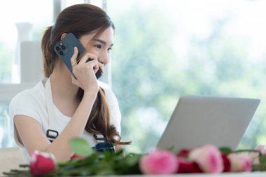 Güzel Asyalı kadın telefonda müşteriyle konuşuyor ve çiçekçide dizüstü bilgisayar kullanıyor..