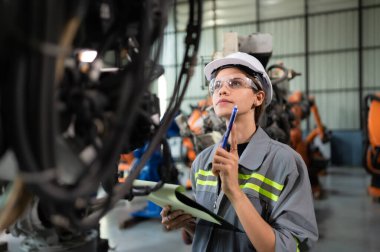 Robot bir depoda robot kola program yükledikten sonra belgelenmiş eşyaları kontrol eden ve operasyonu test eden bir kadın mühendis.