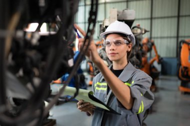 Robot bir depoda robot kola program yükledikten sonra belgelenmiş eşyaları kontrol eden ve operasyonu test eden bir kadın mühendis.