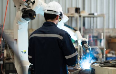 Genç erkek mühendisler kaynak yapan robot eli manipüle ediyorlar. Kaynak manipülasyonunu kaynak denetimi için kontrol paneliyle birleştirerek. Hızlı ve güvenli.