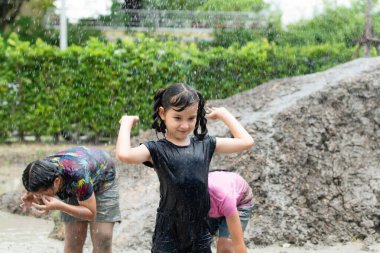 Küçük kızlar toplum sahasında çamurda oynarken eğleniyorlar. Aileler uzaktan izlerken.