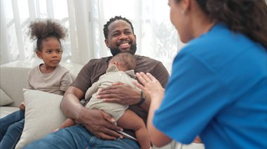 Bir doktor, babasının kollarında uyuyan küçük bir çocuğun sağlığını kontrol etmek için steteskop kullanır.