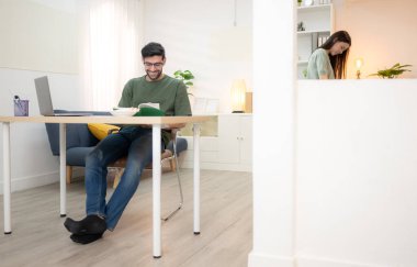 Evde kız arkadaşıyla bilgisayarla çalışırken gülümseyen genç bir serbest çalışanın portresi.