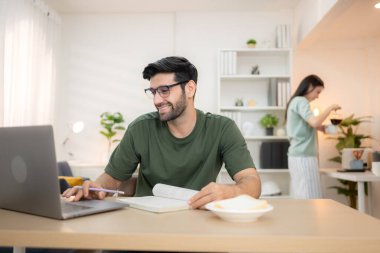 Evde kız arkadaşıyla bilgisayarla çalışırken gülümseyen genç bir serbest çalışanın portresi.