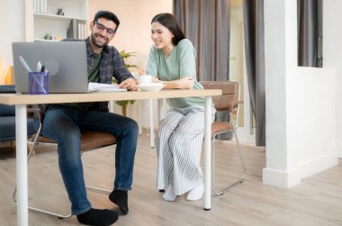 Evde kız arkadaşıyla bilgisayarla çalışırken gülümseyen genç bir serbest çalışanın portresi.