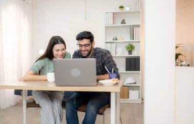 Evde kız arkadaşıyla bilgisayarla çalışırken gülümseyen genç bir serbest çalışanın portresi.