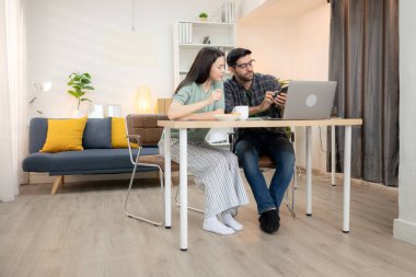 Evde kız arkadaşıyla bilgisayarla çalışırken gülümseyen genç bir serbest çalışanın portresi.