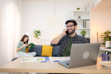 Evde kız arkadaşıyla bilgisayarla çalışırken gülümseyen genç bir serbest çalışanın portresi.