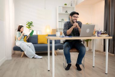 Evde kız arkadaşıyla bilgisayarla çalışırken gülümseyen genç bir serbest çalışanın portresi.