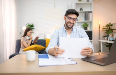 Evde kız arkadaşıyla bilgisayarla çalışırken gülümseyen genç bir serbest çalışanın portresi.