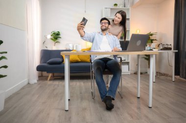 Evde kız arkadaşıyla bilgisayarla çalışırken gülümseyen genç bir serbest çalışanın portresi.