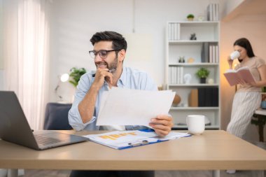 Evde kız arkadaşıyla bilgisayarla çalışırken gülümseyen genç bir serbest çalışanın portresi.