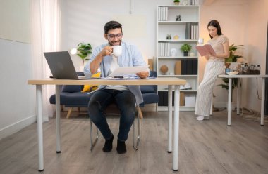 Evde kız arkadaşıyla bilgisayarla çalışırken gülümseyen genç bir serbest çalışanın portresi.