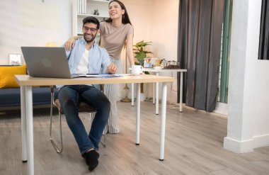 Evde kız arkadaşıyla bilgisayarla çalışırken gülümseyen genç bir serbest çalışanın portresi.