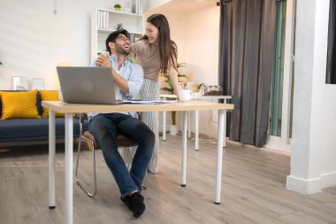 Evde kız arkadaşıyla bilgisayarla çalışırken gülümseyen genç bir serbest çalışanın portresi.