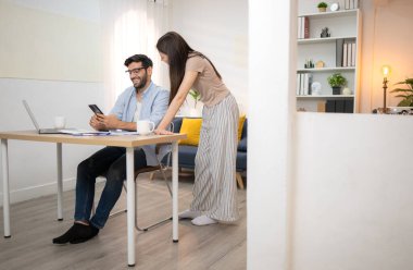 Evde kız arkadaşıyla bilgisayarla çalışırken gülümseyen genç bir serbest çalışanın portresi.