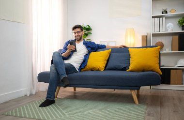 Akıllı telefon kullanan ve evdeki oturma odasında eğlenen genç bir serbest çalışanın portresi.