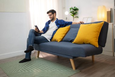 Akıllı telefon kullanan ve evdeki oturma odasında eğlenen genç bir serbest çalışanın portresi.