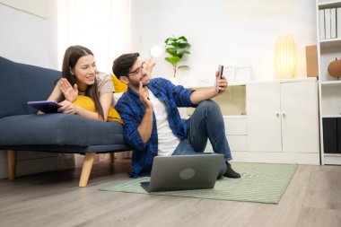 Genç çift rahatlarken gülümsüyor ve evdeki oturma odasında dizüstü bilgisayarla çalışıyorlar.