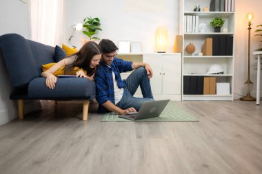 Genç çift rahatlarken gülümsüyor ve evdeki oturma odasında dizüstü bilgisayarla çalışıyorlar.