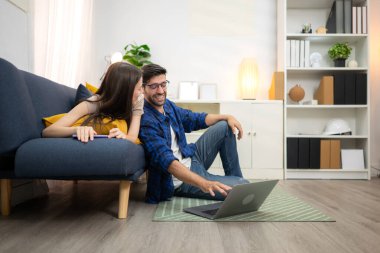 Genç çift rahatlarken gülümsüyor ve evdeki oturma odasında dizüstü bilgisayarla çalışıyorlar.