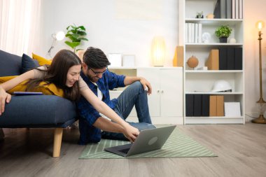 Genç çift rahatlarken gülümsüyor ve evdeki oturma odasında dizüstü bilgisayarla çalışıyorlar.