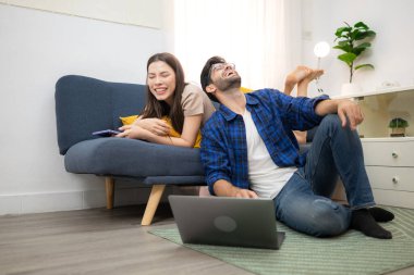 Genç çift rahatlarken gülümsüyor ve evdeki oturma odasında dizüstü bilgisayarla çalışıyorlar.