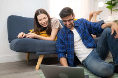 Genç çift rahatlarken gülümsüyor ve evdeki oturma odasında dizüstü bilgisayarla çalışıyorlar.