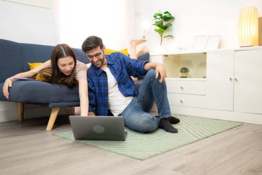 Genç çift rahatlarken gülümsüyor ve evdeki oturma odasında dizüstü bilgisayarla çalışıyorlar.