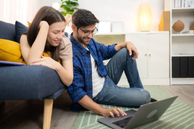 Genç çift rahatlarken gülümsüyor ve evdeki oturma odasında dizüstü bilgisayarla çalışıyorlar.