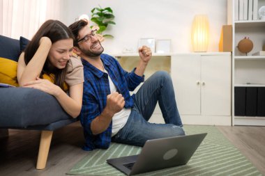 Genç çift rahatlarken gülümsüyor ve evdeki oturma odasında dizüstü bilgisayarla çalışıyorlar.