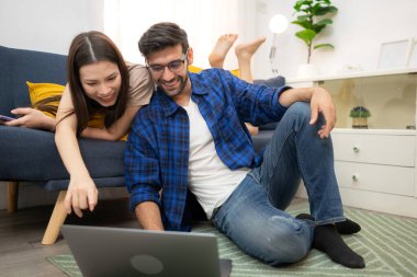 Genç çift rahatlarken gülümsüyor ve evdeki oturma odasında dizüstü bilgisayarla çalışıyorlar.