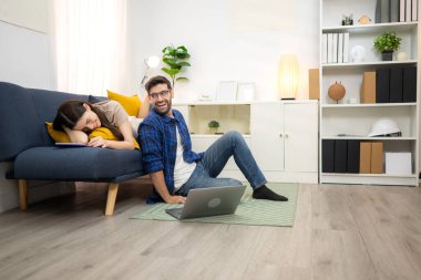 Genç çift rahatlarken gülümsüyor ve evdeki oturma odasında dizüstü bilgisayarla çalışıyorlar.