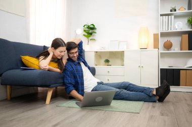 Genç çift rahatlarken gülümsüyor ve evdeki oturma odasında dizüstü bilgisayarla çalışıyorlar.