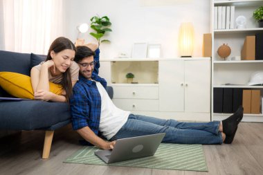 Genç çift rahatlarken gülümsüyor ve evdeki oturma odasında dizüstü bilgisayarla çalışıyorlar.