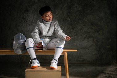 Eskrim kıyafeti giymiş genç bir çocuk bankta oturmuş kılıcını dikkatle inceliyor. Yanına yerleştirilmiş miğfer.