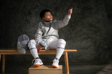 Eskrim kıyafeti giymiş genç bir çocuk bankta oturmuş kılıcını dikkatle inceliyor. Yanına yerleştirilmiş miğfer.