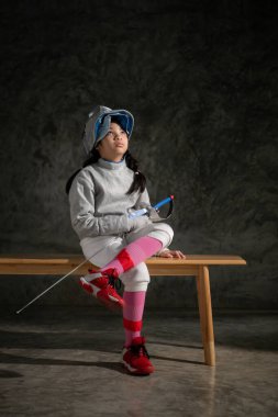 Eskrim kıyafetli bir kız bankta oturuyor, hazırlık yarışmasına hazır.