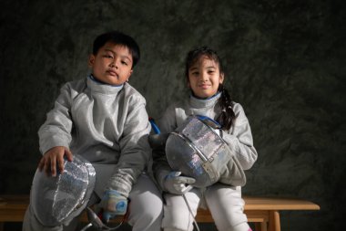 Eskrim kıyafeti giymiş iki çocuk bir bankta oturup birbirlerine gülümsüyor. Maskelerini ve kılıçlarını tutarlar. Eğitimden önce veya sonra bir anlarını paylaşırlar.