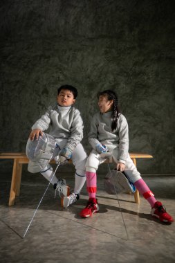Eskrim kıyafeti giymiş iki çocuk bir bankta oturup birbirlerine gülümsüyor. Maskelerini ve kılıçlarını tutarlar. Eğitimden önce veya sonra bir anlarını paylaşırlar.