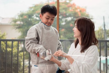 Eskrim kıyafeti giymiş bir çocuk anneleriyle birlikte yaklaşan müsabaka gününe hazırlanmak için eskrim antrenmanından önce yiyecek ve spor malzemeleri hazırlıyor.