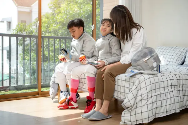 Bir kız ve oğlan, anneleriyle eskrim kıyafetleri içinde yaklaşan müsabakaya hazırlanmak için eskrim antrenmanından önce yiyecek ve spor malzemeleri hazırlıyorlar.