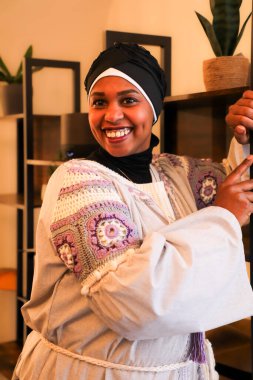 Etnik beyaz boho tarzında gamzeli siyahi bir kadın. İçerde büyüleyici bir şekilde gülümsüyor. Raflara yaslanıyor.