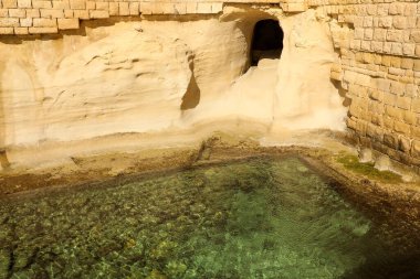 On the Maltese rocky coast, next to the turquoise transparent water of the sea, in the corner of the fortress walls, there is a secret and inconspicuous grotto. clipart