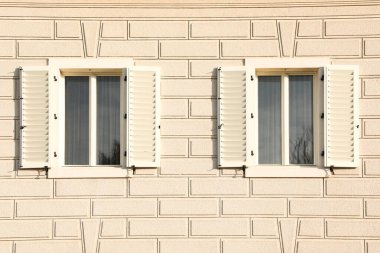 Avrupa şehirlerinden birinde güneşli bir günde beyaz bir cephede tuğla taklidi yapan bir evde panjurları olan iki şık pencere..