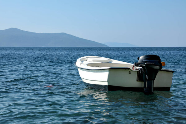 Small white motorboat quietly floats on clear blue sea water, with distant mountains under a cloudless sky, capturing the serenity of summer travel and coastal adventure.
