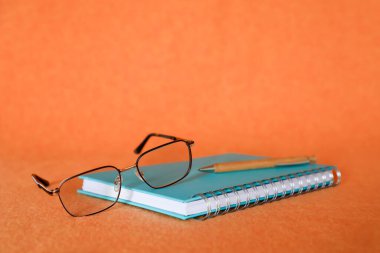 Okuma gözlüğü ve şık bir tükenmez kalem zarif bir şekilde spiral bir deftere oturur, parlak turuncu bir zemin ile vurgulanır, bilgiyi, öğrenmeyi ve entelektüel gelişimi simgeler..