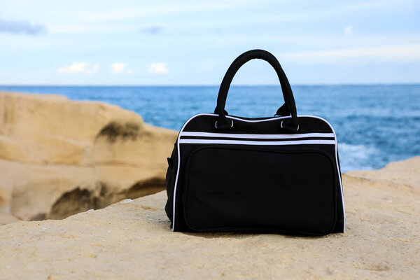Sleek black travel bag sits on a rocky shoreline by the sea, captured up close. The scene highlights modern style, a spirit of adventure, and the atmosphere of coastal travel.