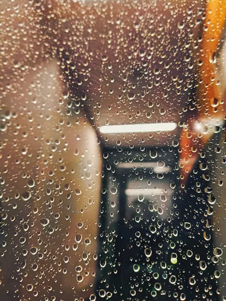 Pencereden sarkan yağmur damlaları arka planda bulanık ışıkla yumuşak, dokulu bir etki yaratıyor. Geçen manzaranın bulanık turuncu ve gri tonlarını yansıtan.